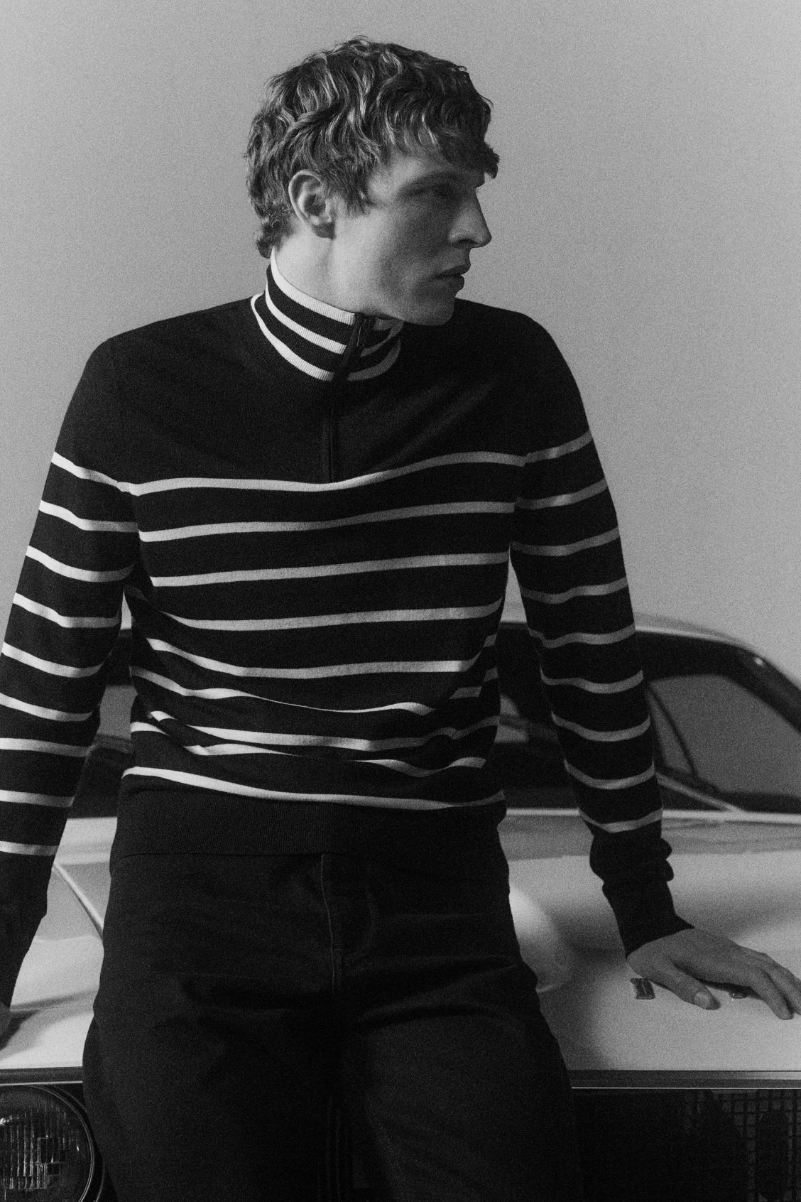 Young man sitting on a car hood, wearing black jeans and a striped COS sweater.