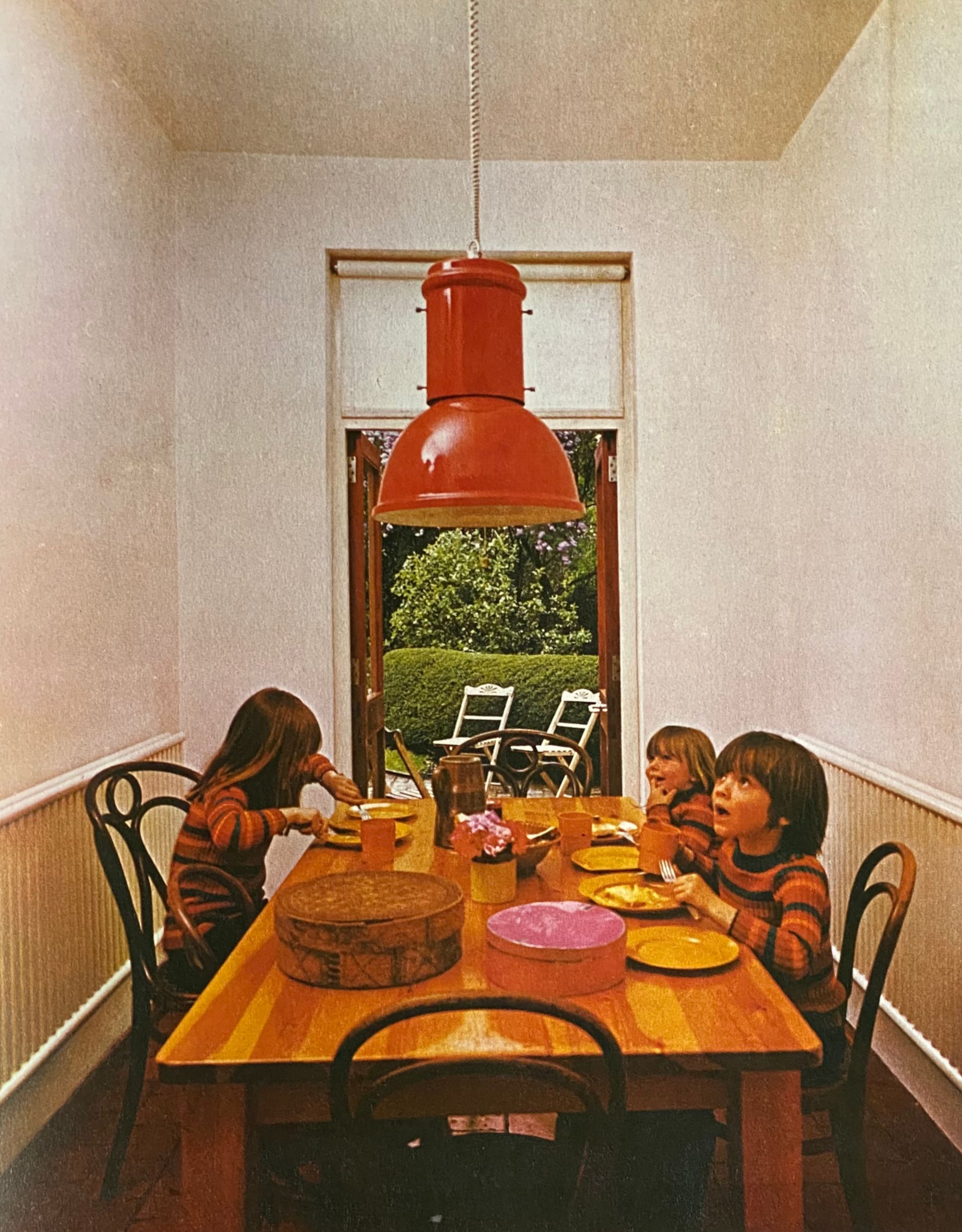 Three children eating at a wooden dining room table with a large red industrial lamp suspended above it.