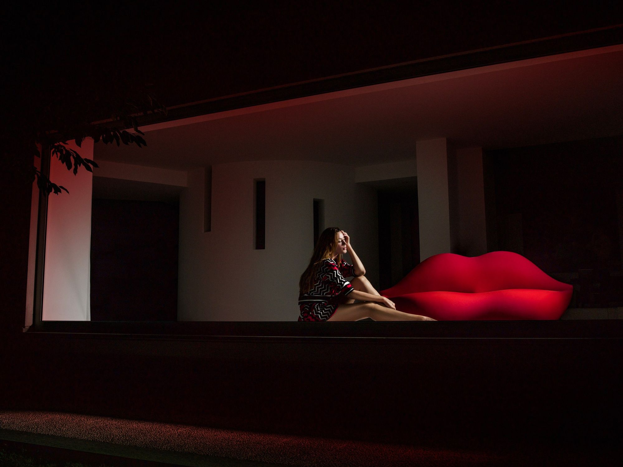 Bright red Bocca lips sofa in a dimly lit room with a pensive girl sitting on the floor nearby