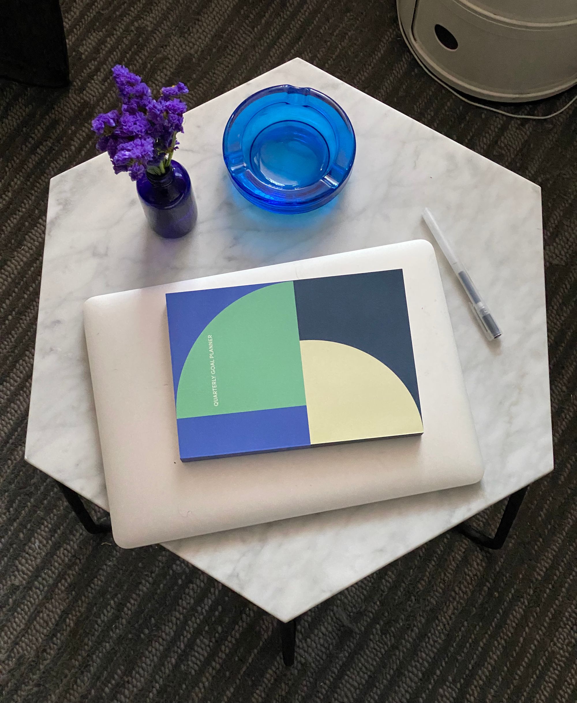 Hexagonal marble tabletop seen from above, with laptop, notebook, pen, ashtray, and bud vase on it.