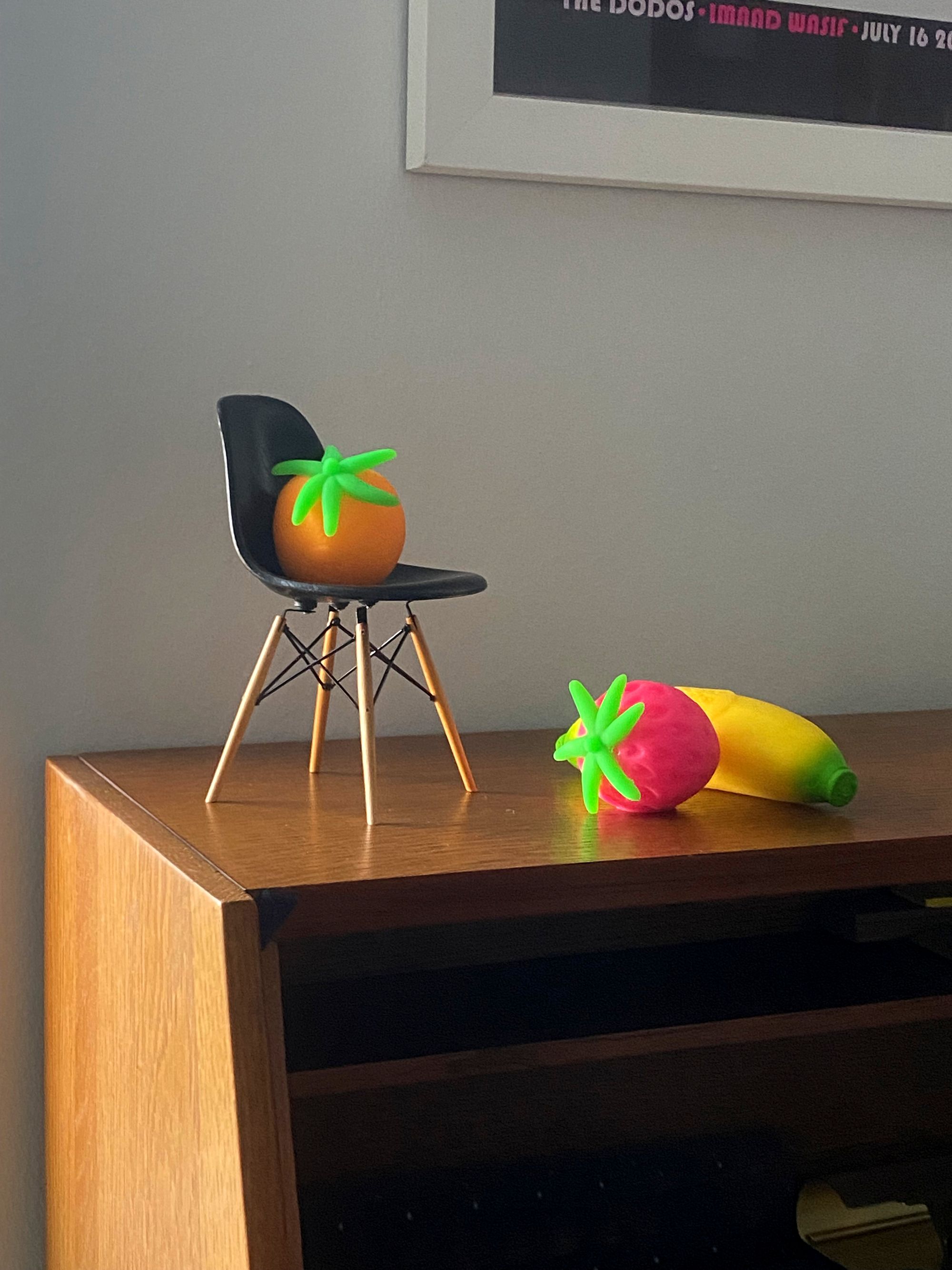 Miniature Eames side chair holding an orange-shaped stress toy, with strawberry and banana stress toys resting nearby.