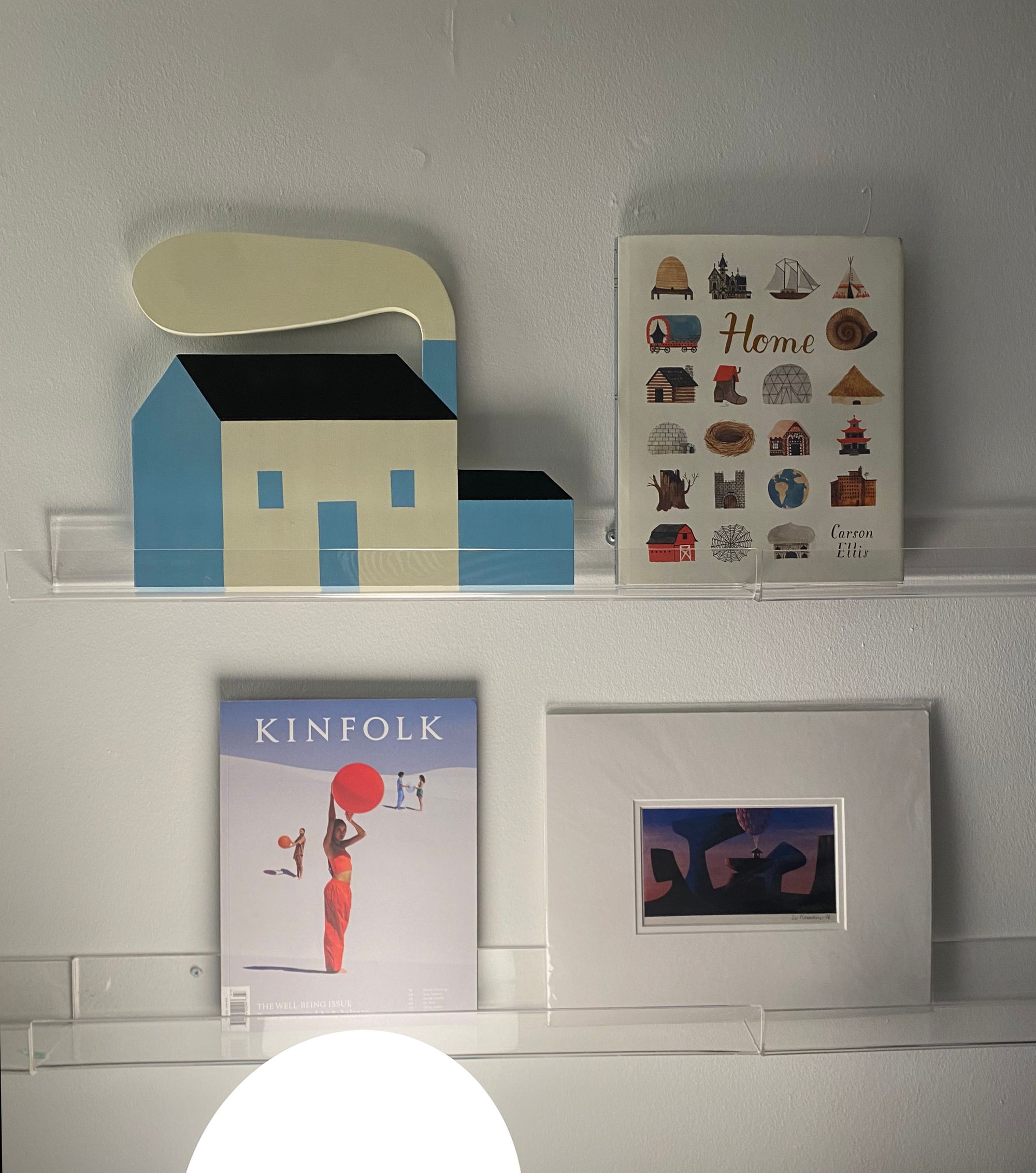 Two clear acrylic wall ledges holding an artwork by Erin McPhee, a book, a magazine, and a print.