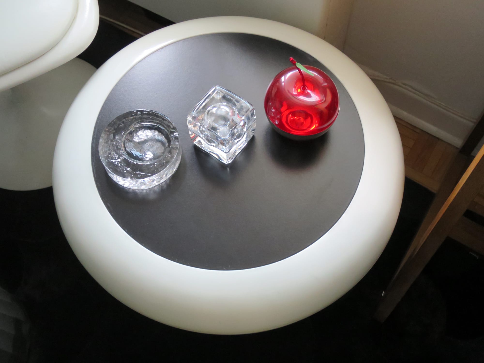 Three decorative objects on a side table: two ashtrays made of clear glass, and a bright red box in the shape of an oversized cherry.