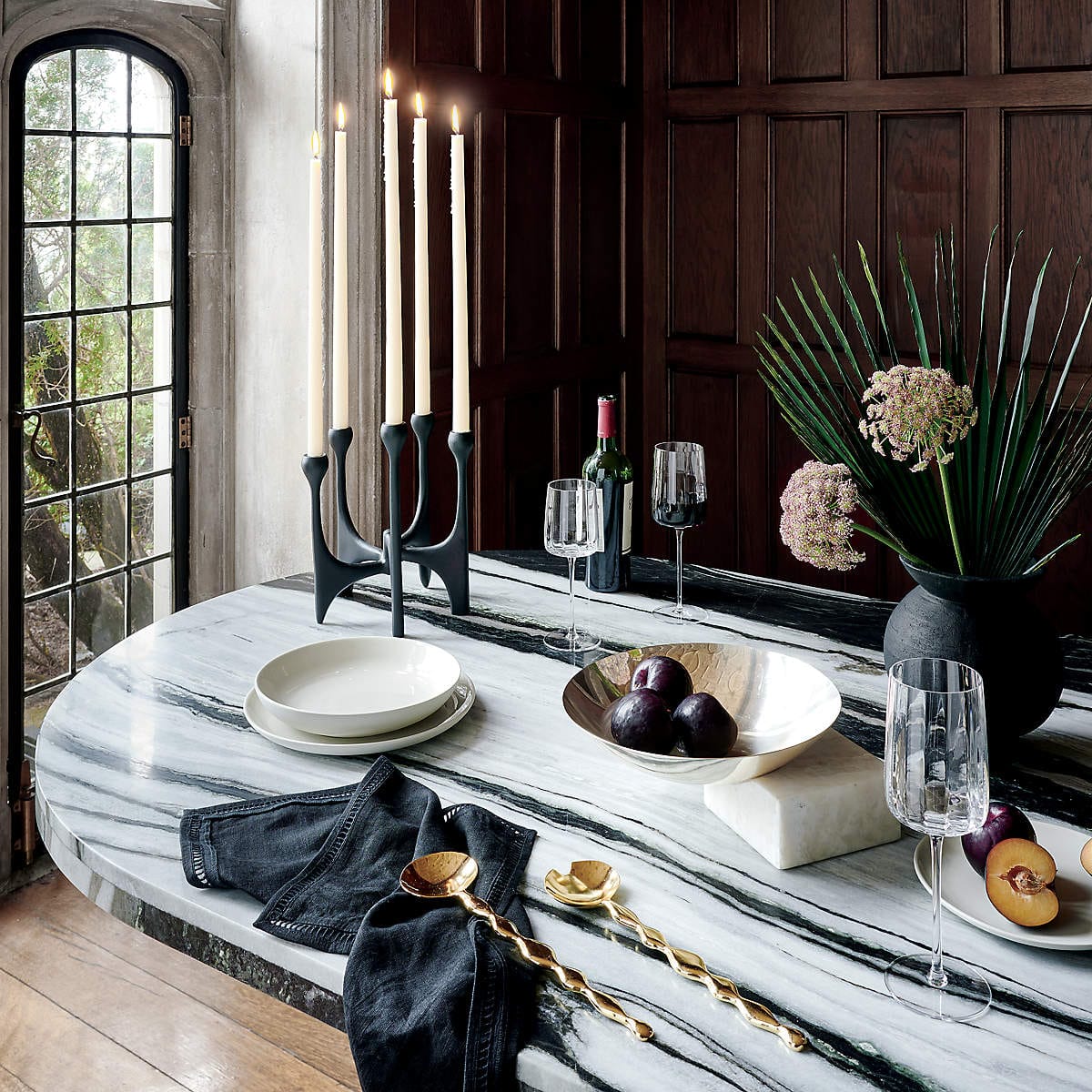 The same black candelabra as above, with five lit white candles, on a marble tabletop with artfully arranged objects.