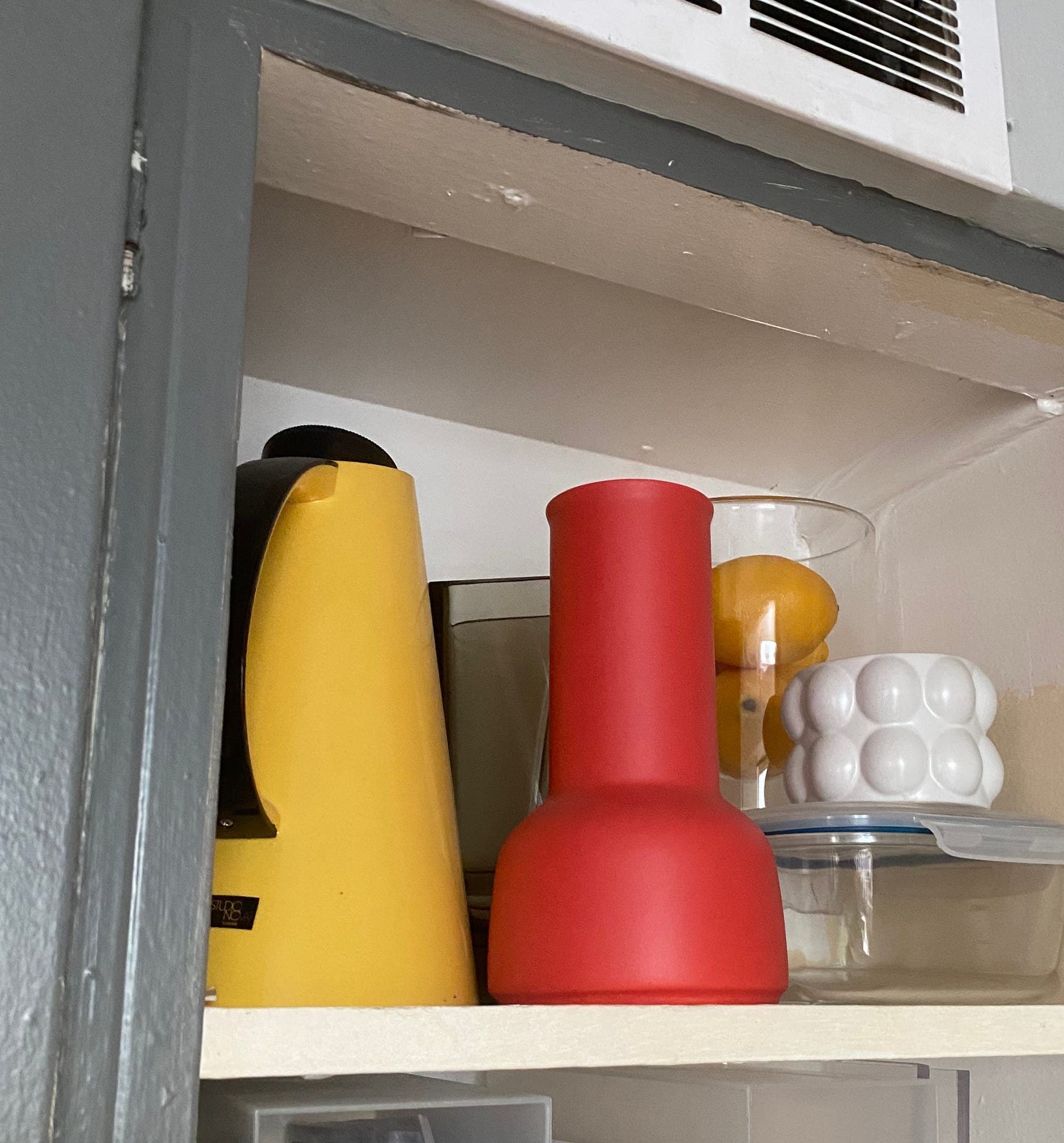 Kitchen cupboard shelf with small objects on it, including a bright red vase and bright yellow pitcher.