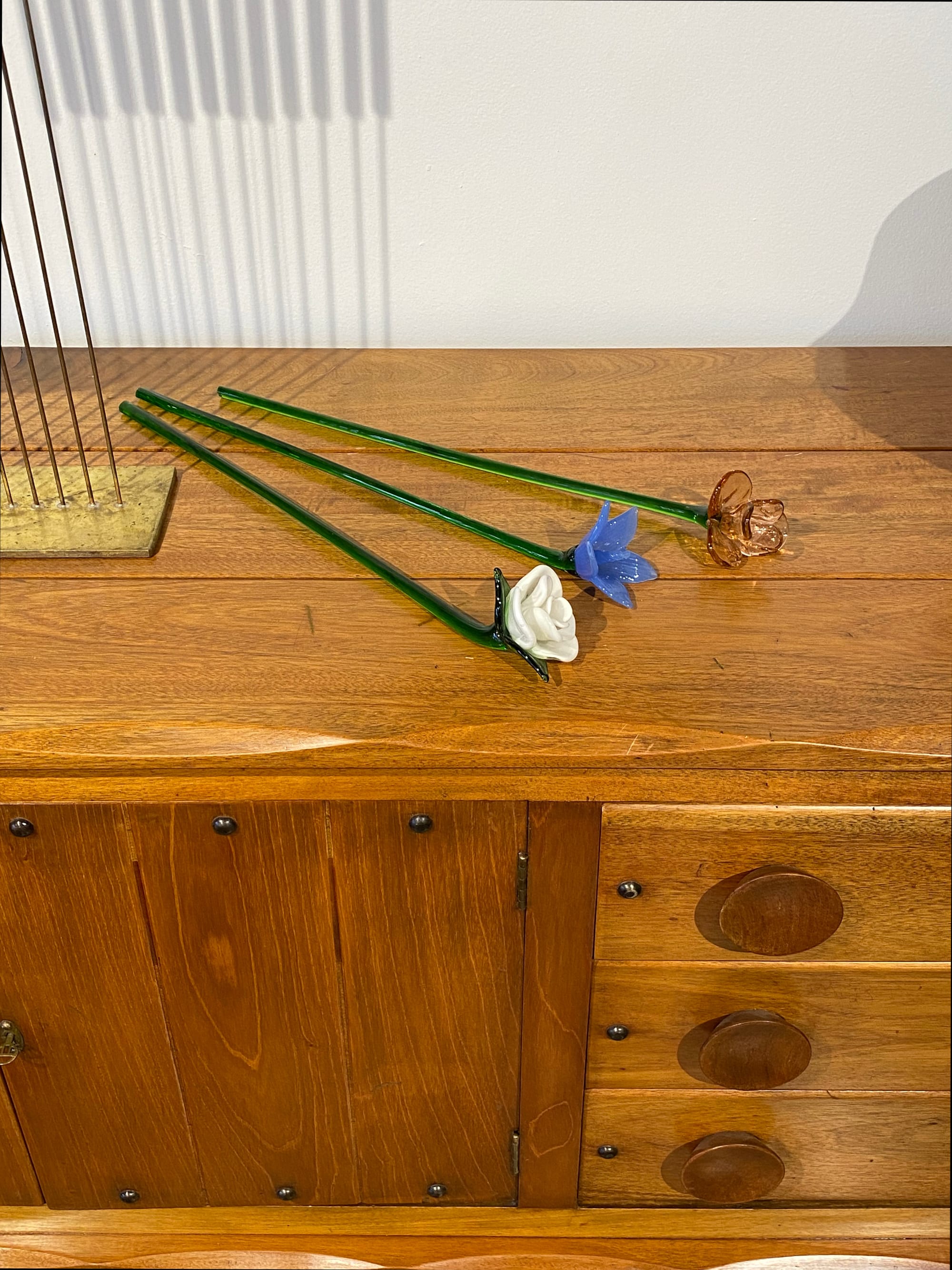 Three glass flowers resting horizontally on a wooden credenza.