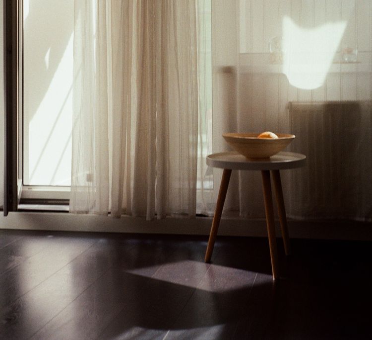 Dimly lit interior featuring a three-legged stool that supports a bowl with a single orange inside.