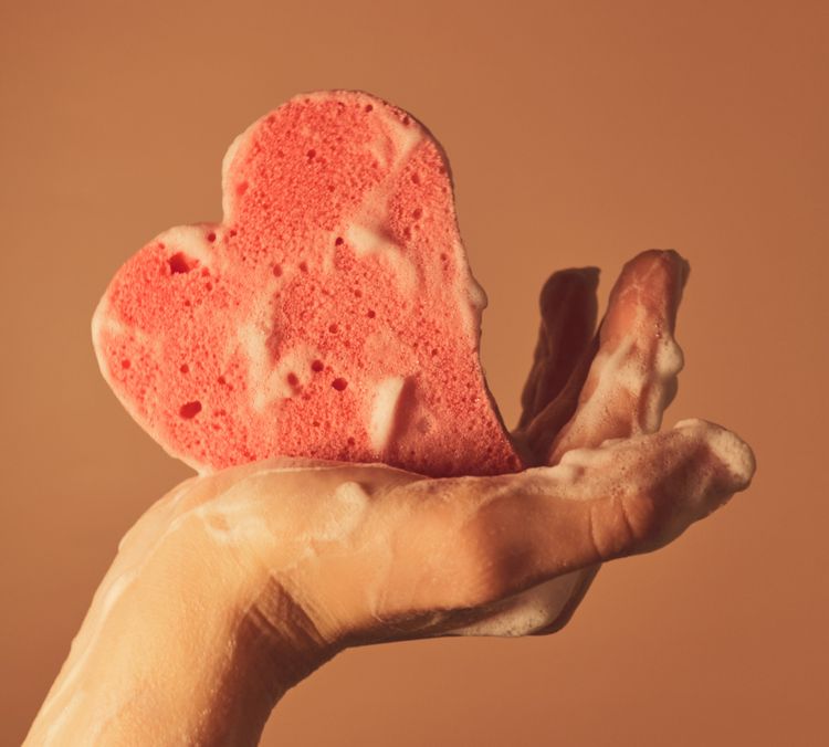 Hand holding a heart-shaped sponge, both covered in lather.