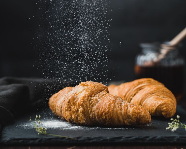 Two croissants against a black backdrop, the one in the foreground with a rain of icing sugar coming down on it.