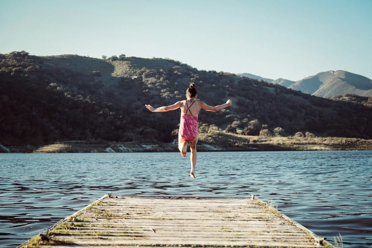 Girl in a pink bathing suit caught in mid-air as she leaps off a dock into a lake.