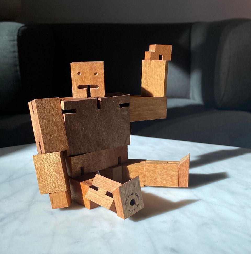 Wooden robot sitting on a marble tabletop with his hand in a waving gesture.