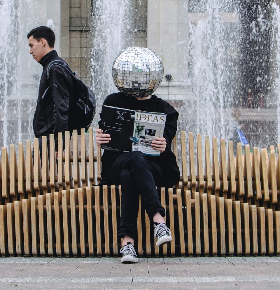 Figure sitting on a bench and holding a magazine called Ideas, their head inside a disco ball.