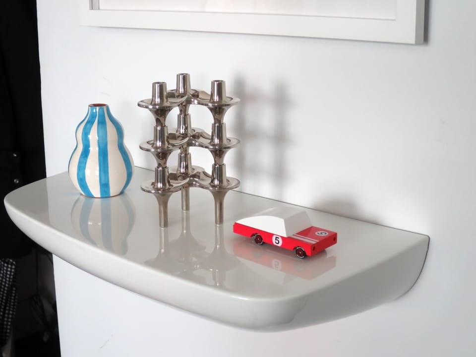 Wall shelf with three items on it: a small vase, a stacked set of chrome candleholders, and a toy car.