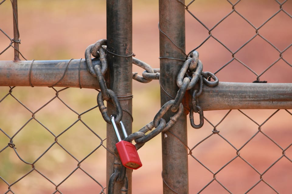 A chain in the shape of a heart, fastened by a red lock, and holding two sections of a fence together.